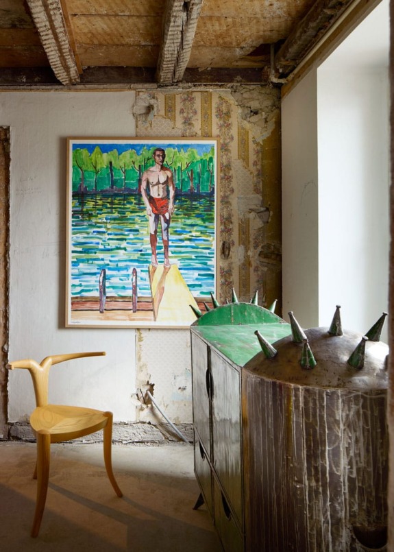 Interior with a modern wooden chair, a spiked metallic storage piece, and a colorful lakeside painting on a textured wall.