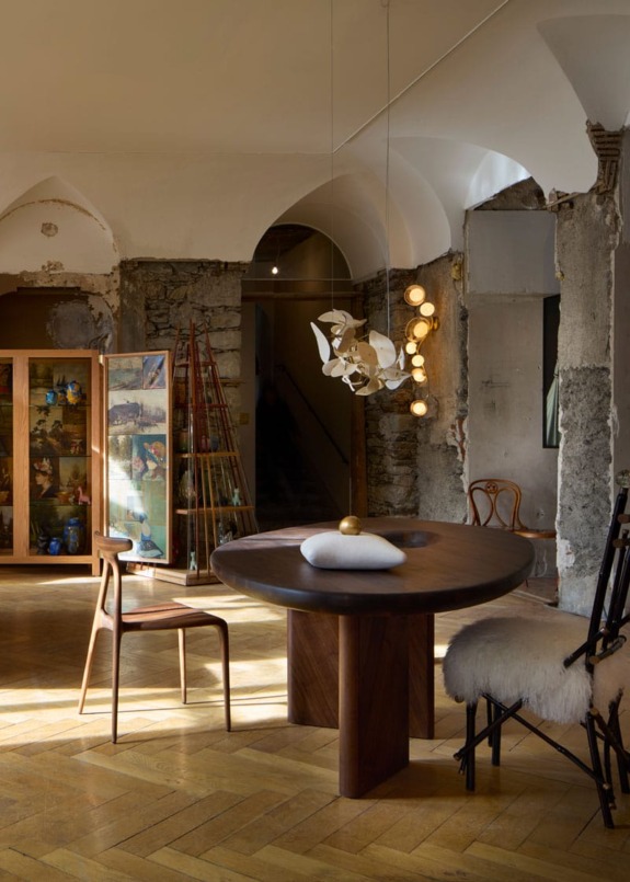 Rustic dining room with wooden table, chairs, modern pendant light, and exposed stone walls in an eclectic interior setting.