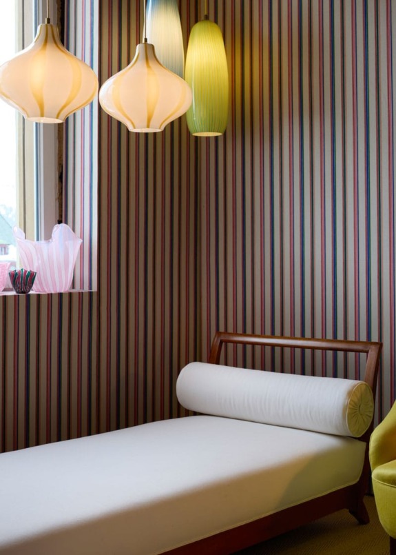 Minimalistic bedroom with striped wallpaper, wooden bed, white cushion, modern pendant lights, and a window with colorful decor.