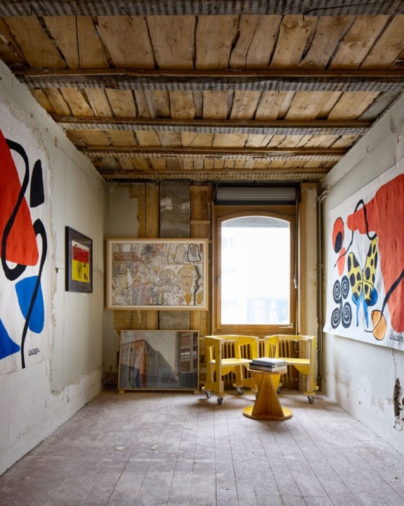 Art studio interior with abstract paintings on walls, wooden ceiling, and a central window filtered with natural light.