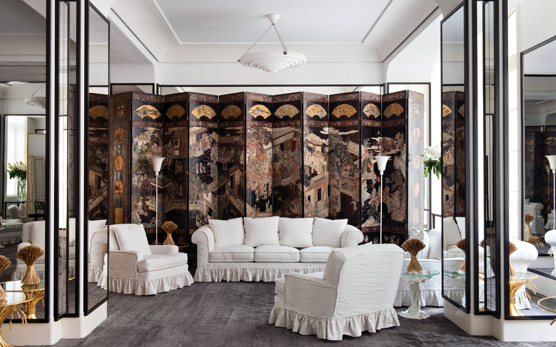 Elegant living room with white sofa and chairs, ornate folding screen, mirrored walls, and modern chandelier.