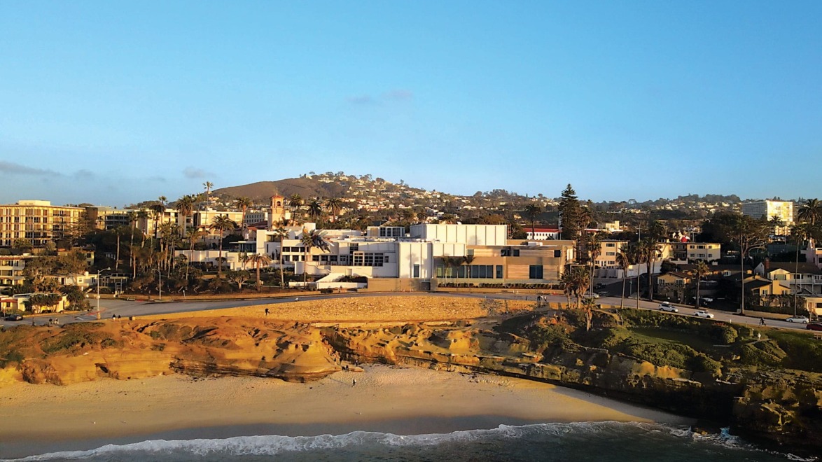 Aerial view of a coastal cityscape with buildings, sandy beach, and ocean under a clear blue sky.