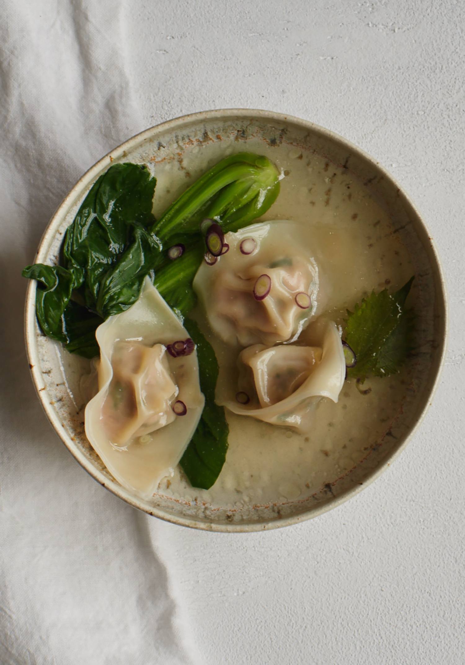 Bowl of soup with dumplings, leafy greens, and garnished with sliced onions on a textured white surface.