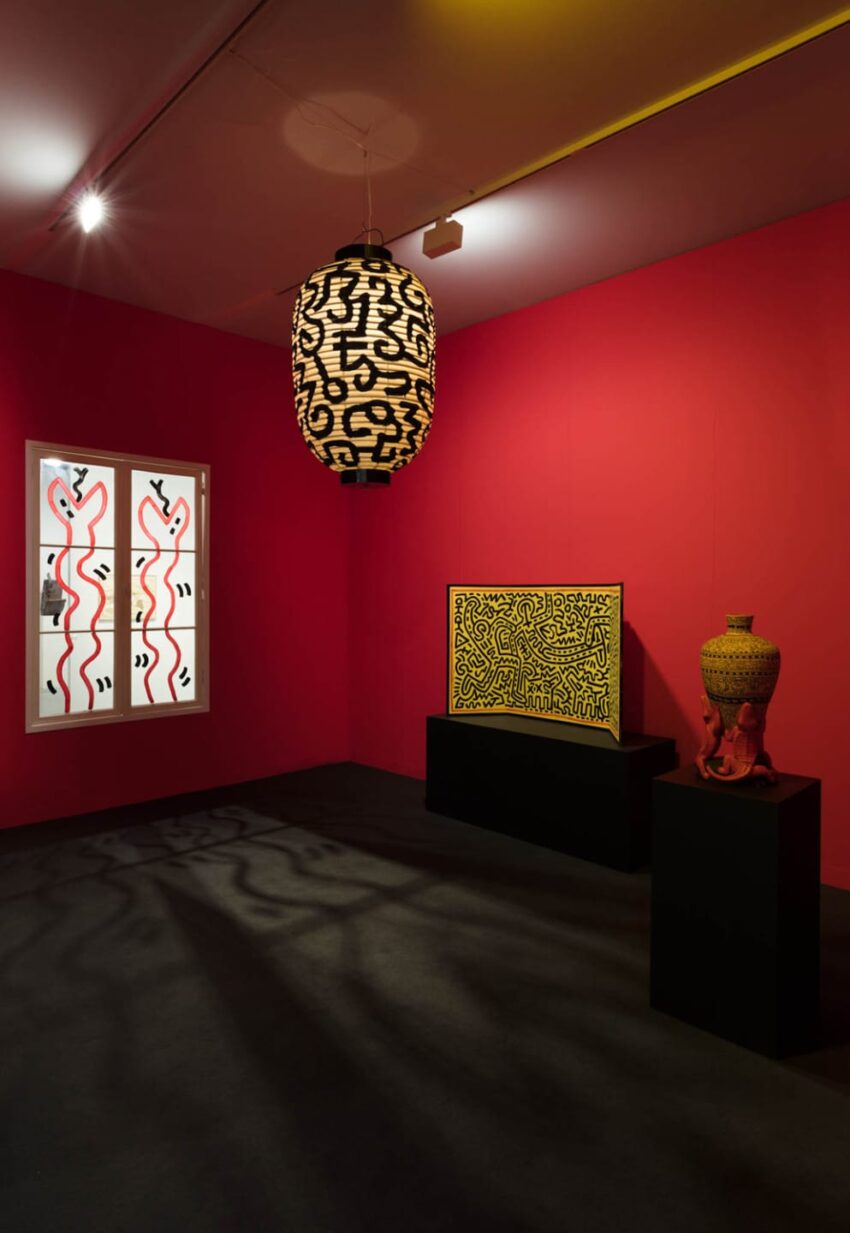 Art gallery with red walls displaying abstract patterned artwork, a decorative lamp, and a vase on pedestals.