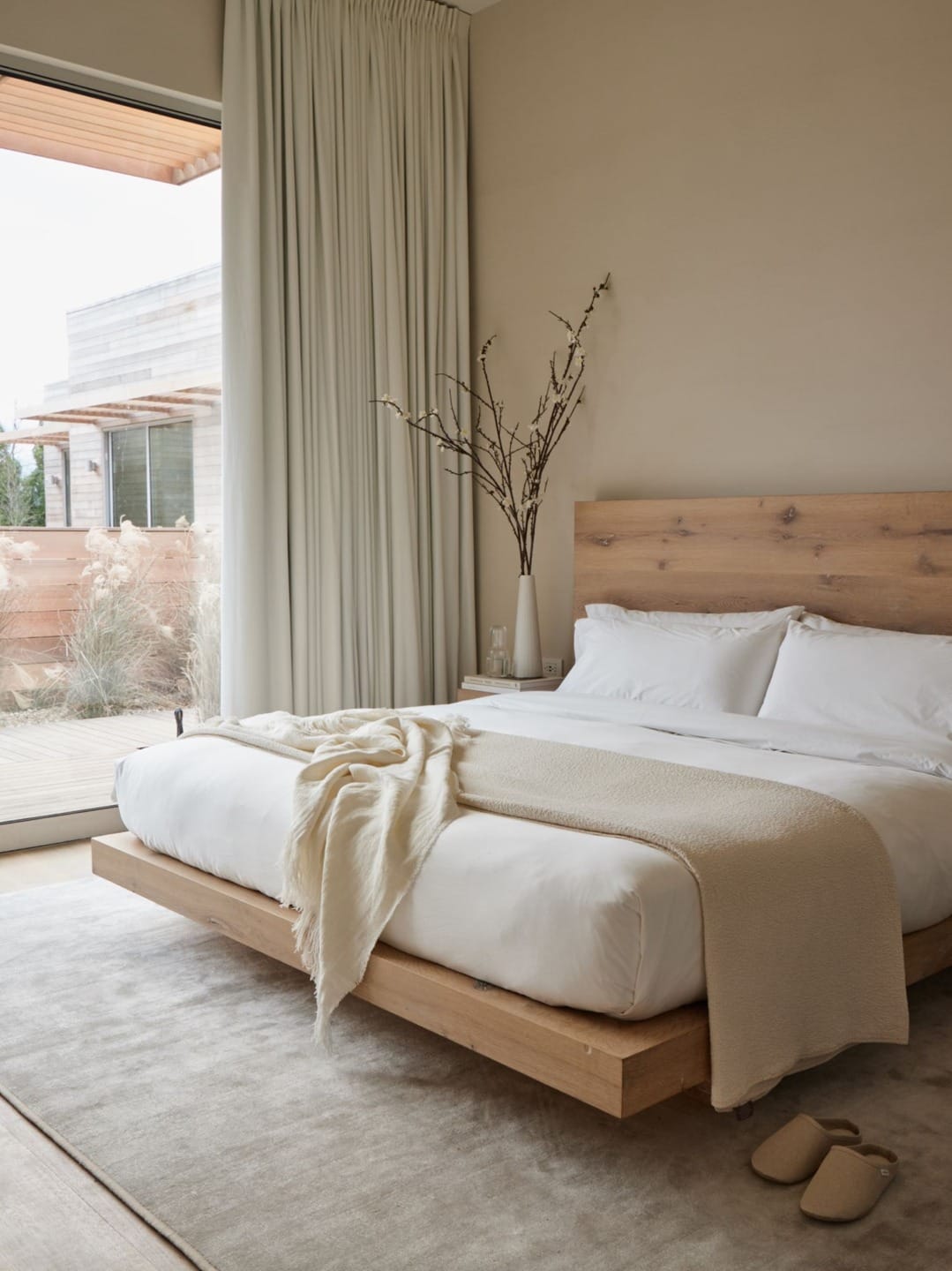 Minimalist bedroom with wooden bed frame, white bedding, beige throw, large window, and light curtains.