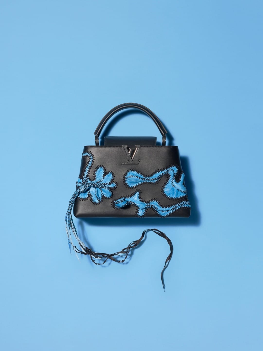 Black handbag with blue floral embroidery and tassels on a light blue background.