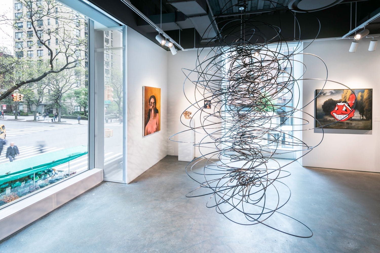 Art gallery interior with abstract wire sculpture in foreground and paintings on walls, large windows showing city street outside.