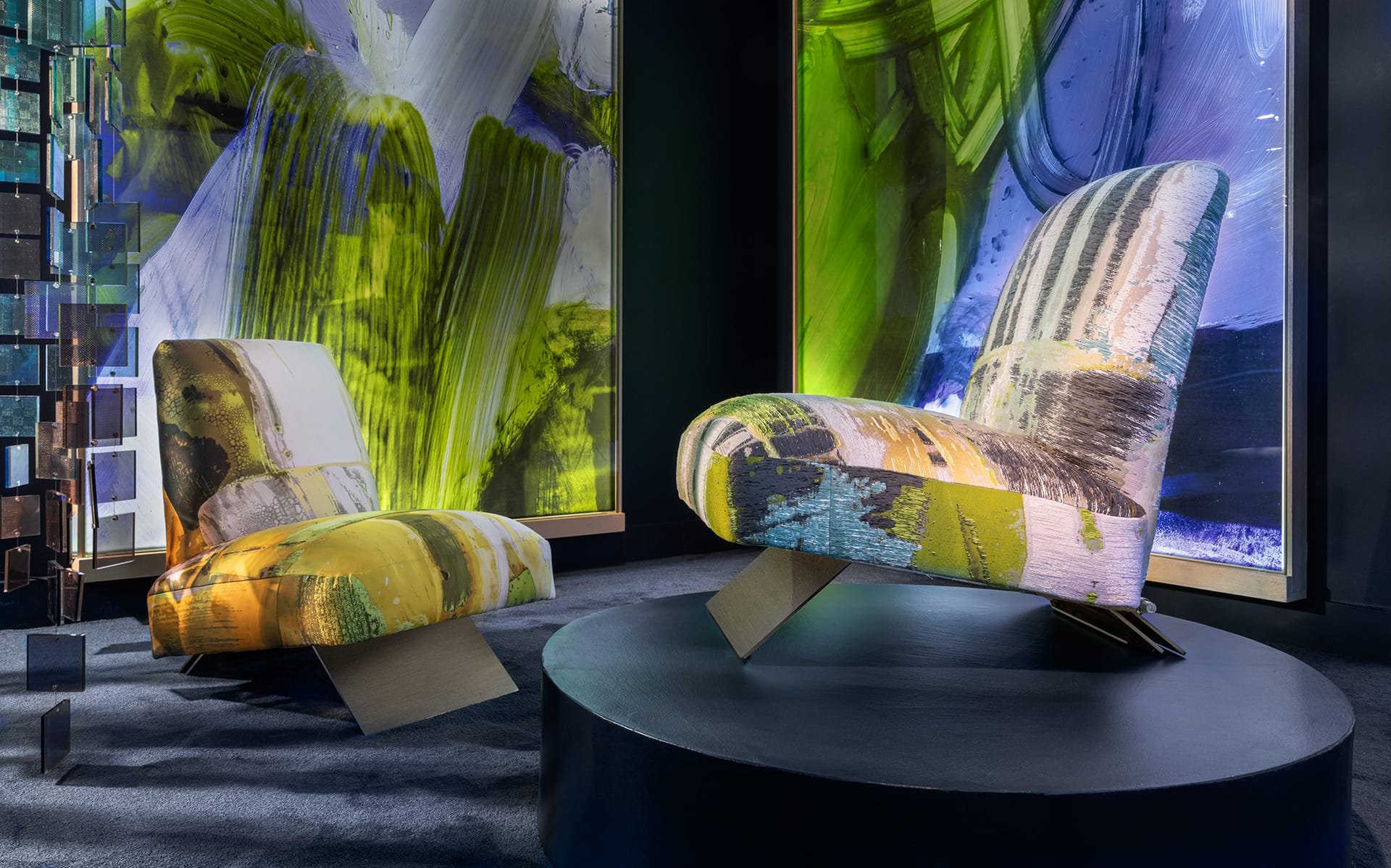 Two modern, abstract-patterned armchairs on a black platform with colorful, artistic backdrop in a gallery setting.