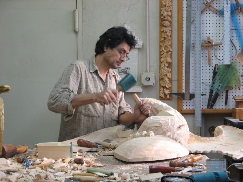 Man carving wood in a workshop filled with tools on the wall and workbench.