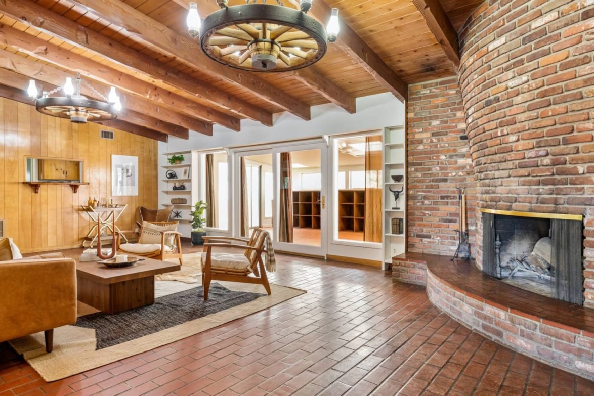 Rustic living room with wooden beams, brick fireplace, leather chairs, and wagon wheel chandeliers. Cozy and inviting space.
