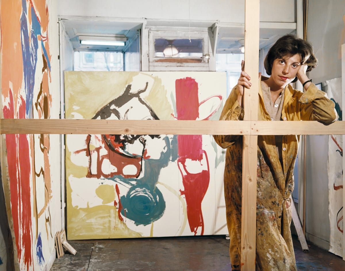 Artist standing in a paint-splattered studio holding a wooden frame, surrounded by colorful abstract paintings.