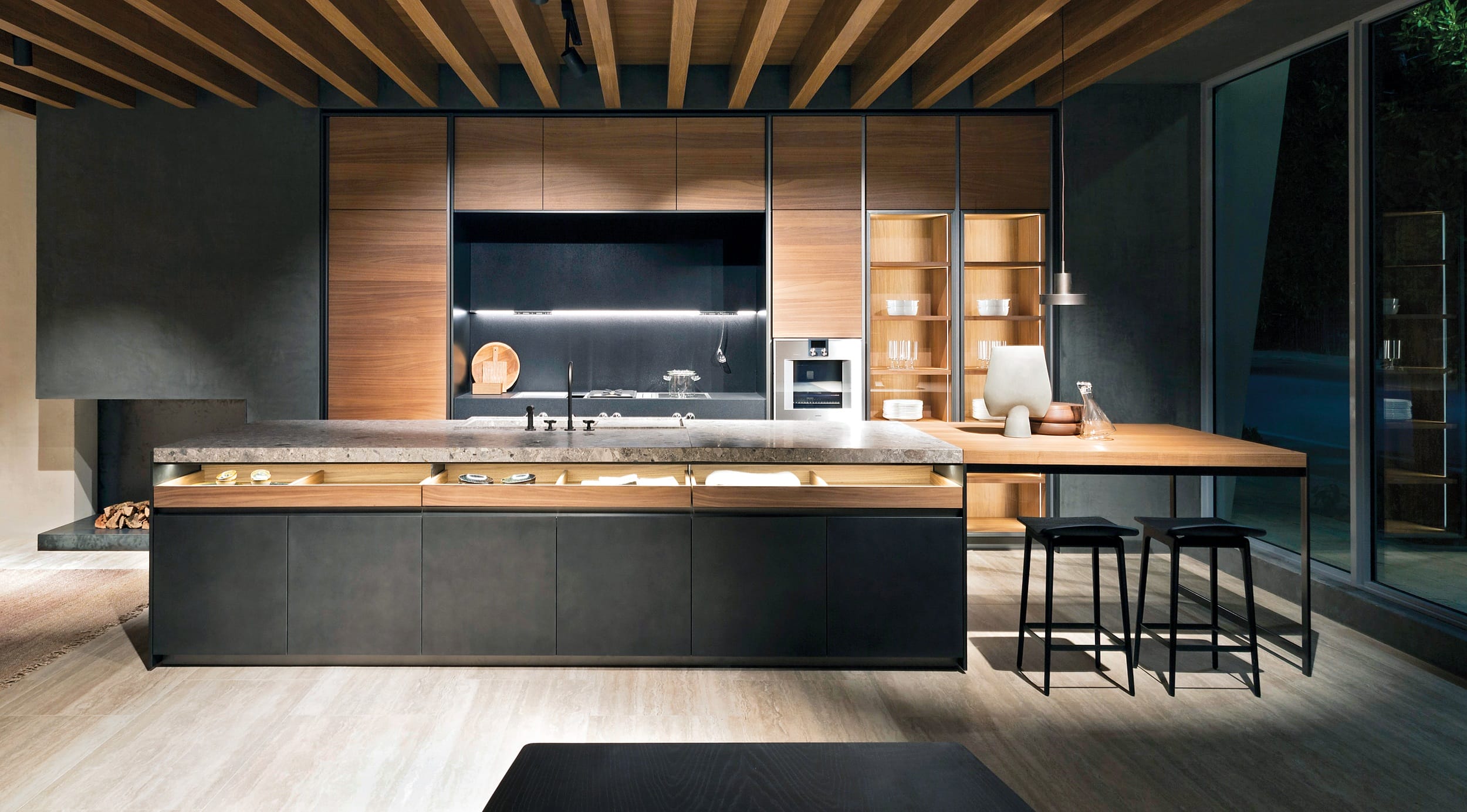 Modern kitchen interior with wood cabinets, island, and stools under a beamed ceiling, showcasing contemporary design.