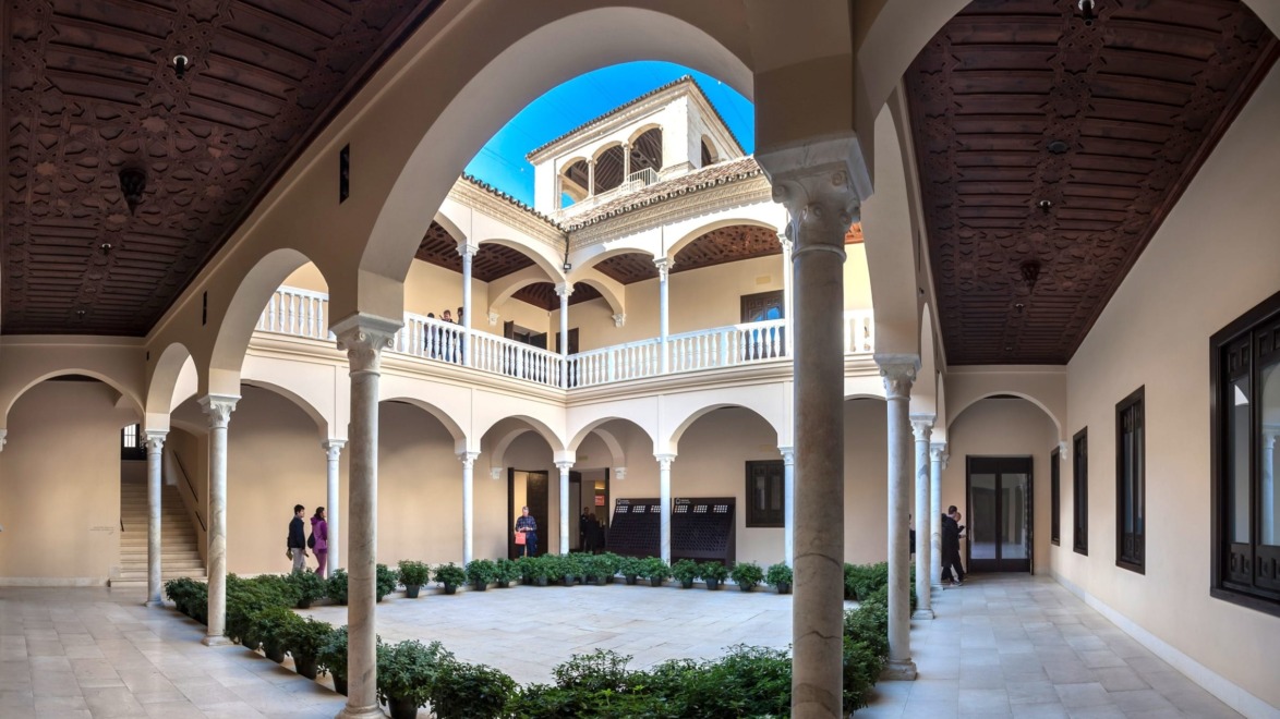 Elegant courtyard with arched columns and lush greenery, featuring a sunny sky and people walking in the background.
