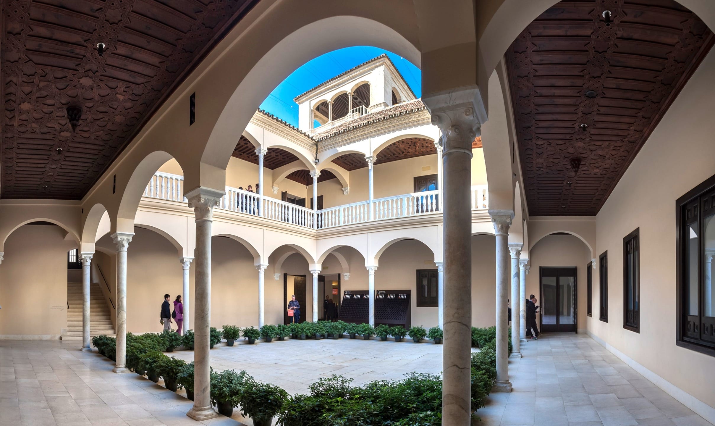 Elegant courtyard with arched columns and lush greenery, featuring a sunny sky and people walking in the background.