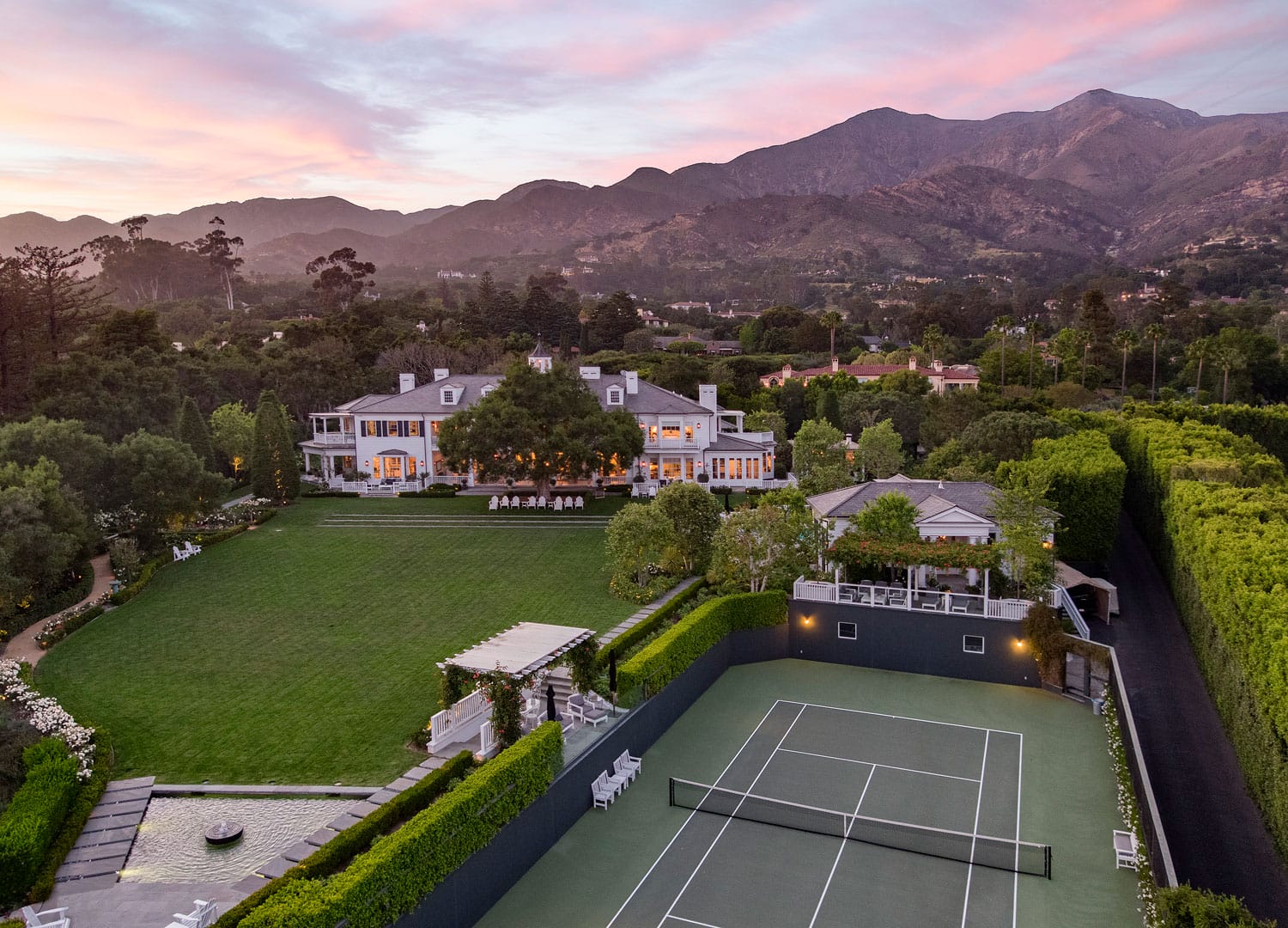 Aerial view of a large estate with a tennis court, lush green lawns, and mountains in the background at sunset.