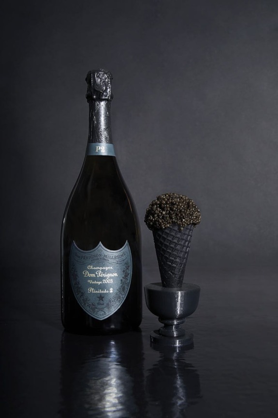 Champagne bottle beside a black ice cream cone topped with caviar on a dark reflective surface.