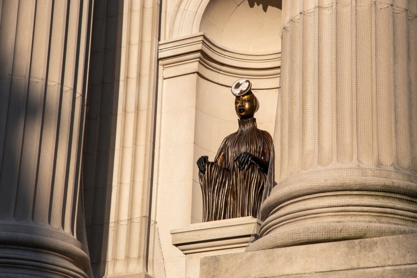 Statue of a seated figure in flowing attire with a modern, circular headpiece in an architectural niche.