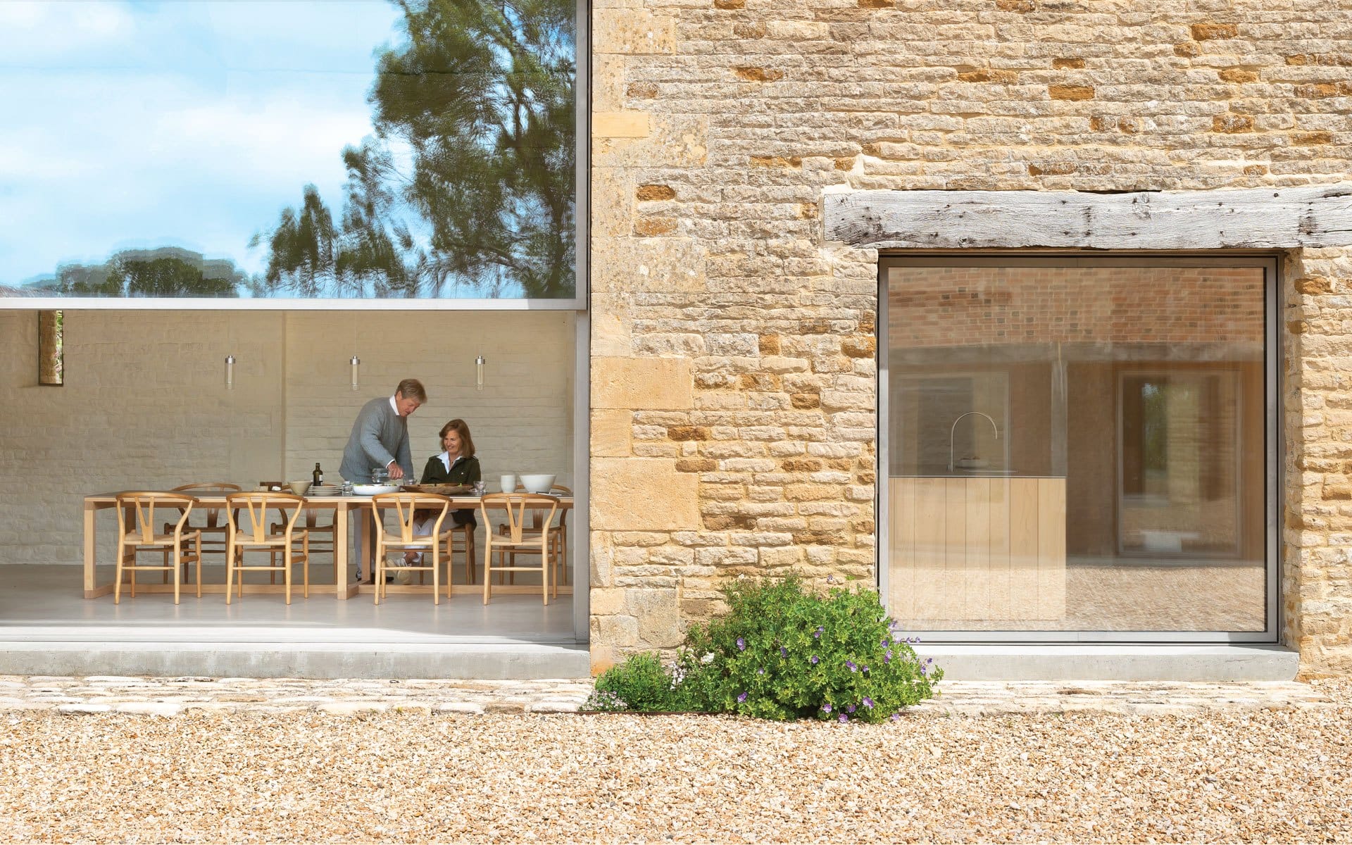 Two people working at a wooden table near a large window in a modern stone house with another window to the right.