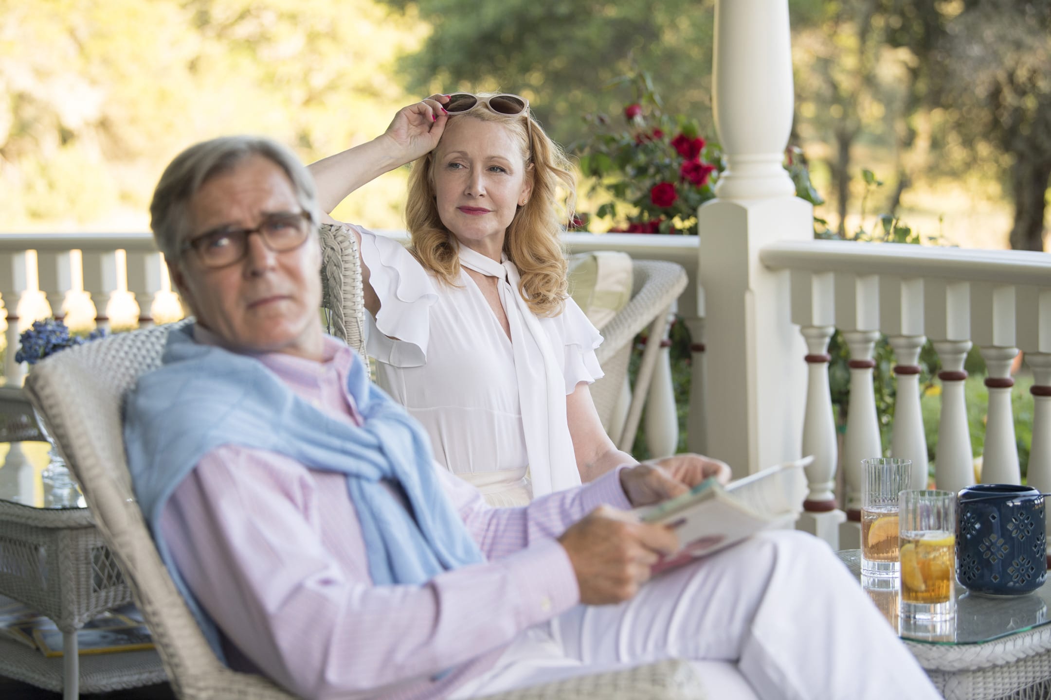 Elderly couple relaxing on a porch with drinks and a newspaper, surrounded by greenery and enjoying a sunny day.