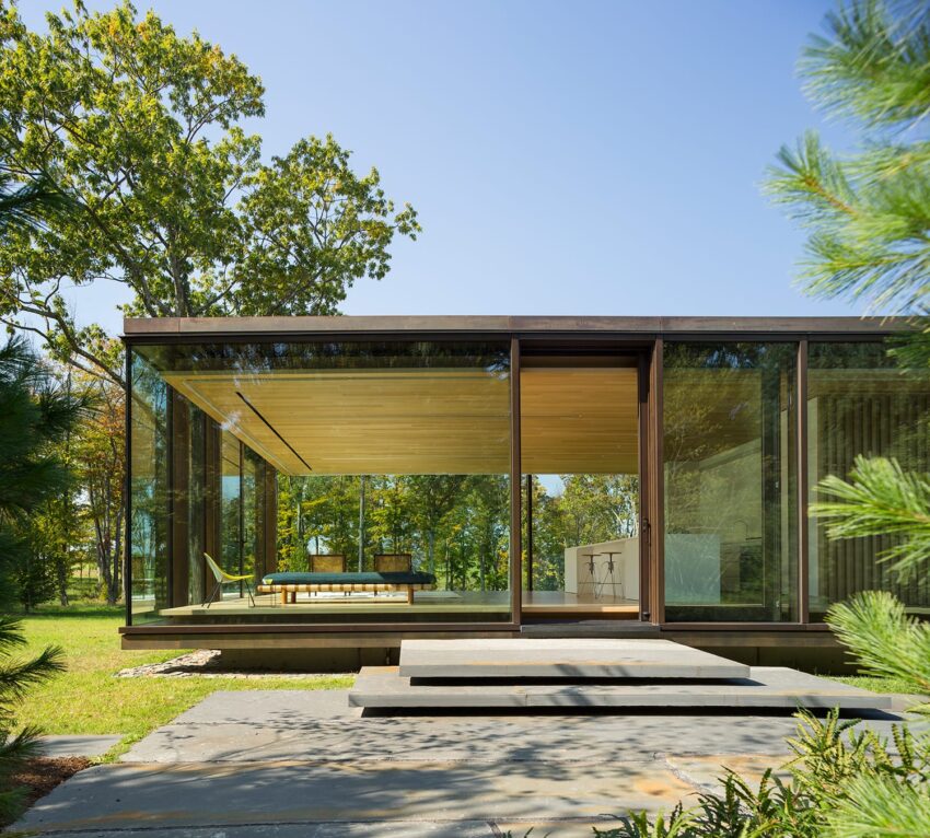 Modern glass house surrounded by trees with a minimalist interior and a view of the outdoors.