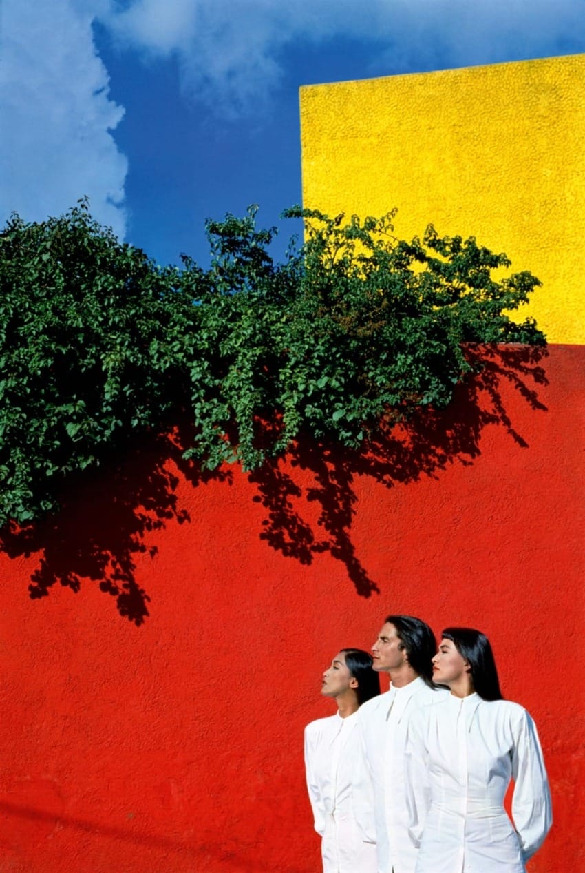 Three people in white outfits stand against a vibrant red and yellow wall with green foliage under a bright blue sky.