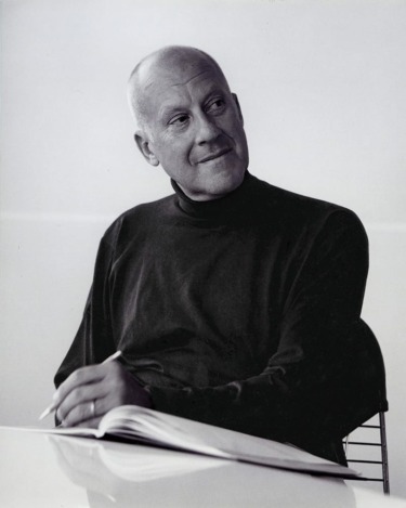 Man in a dark turtleneck sitting while holding a pen and looking thoughtfully to the side with a notebook on the table.
