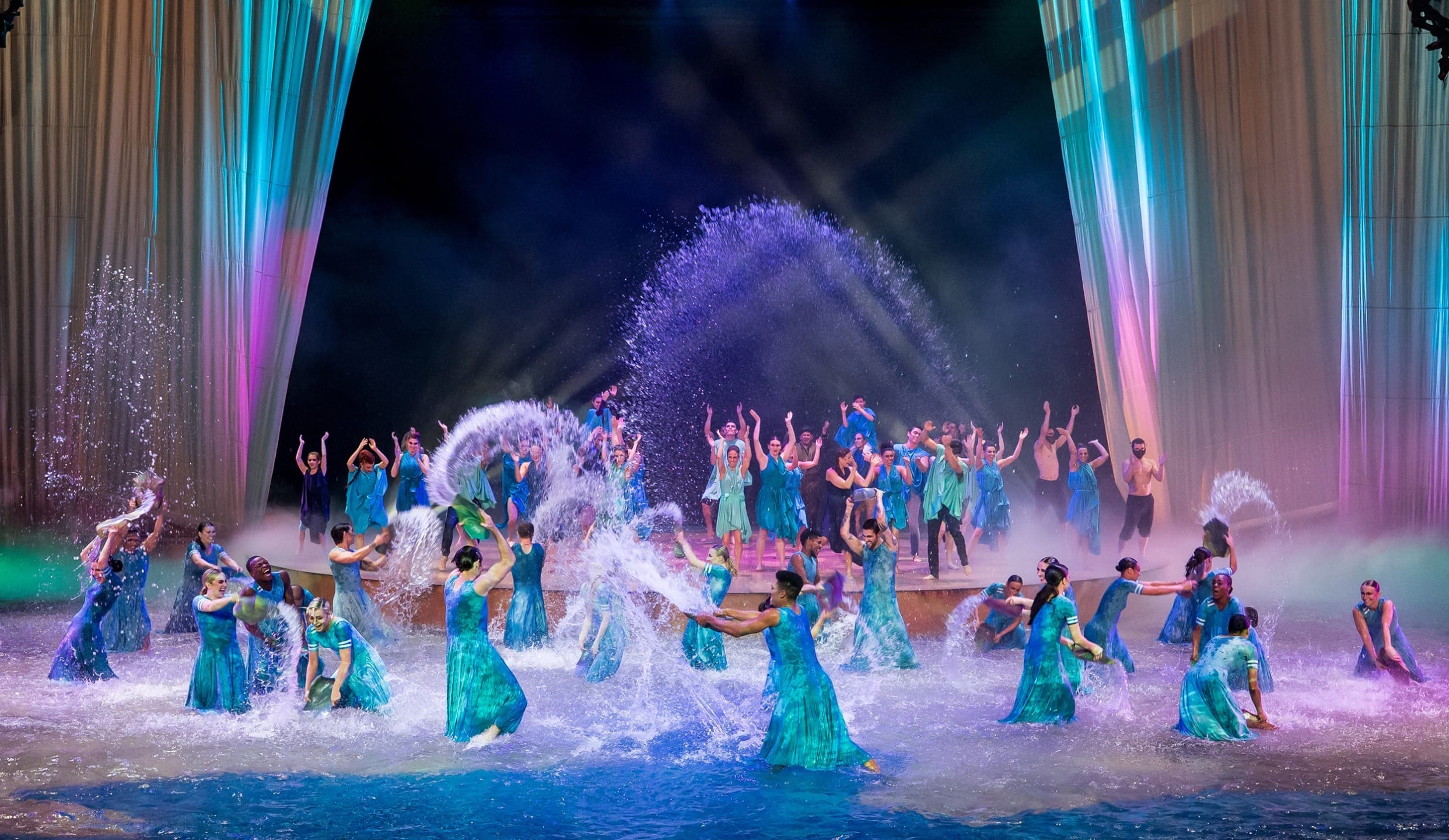 Performers in blue costumes dance in a colorful water show, splashing water under vibrant stage lights.
