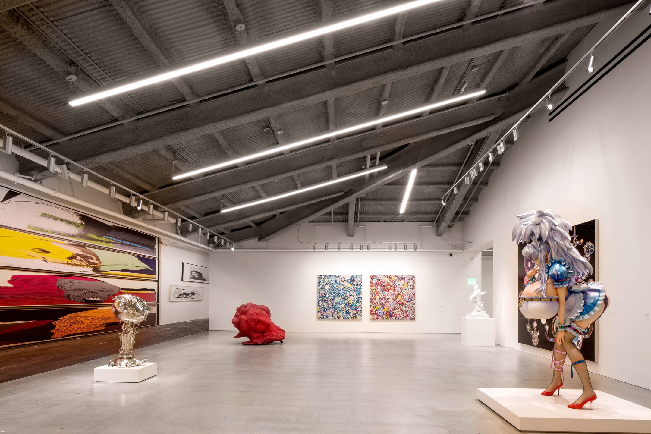 Modern art gallery with various sculptures and colorful paintings displayed in a spacious room with high ceilings.