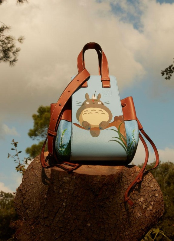 A brown leather tote bag featuring a whimsical cartoon character design, placed on a tree stump against a cloudy sky.