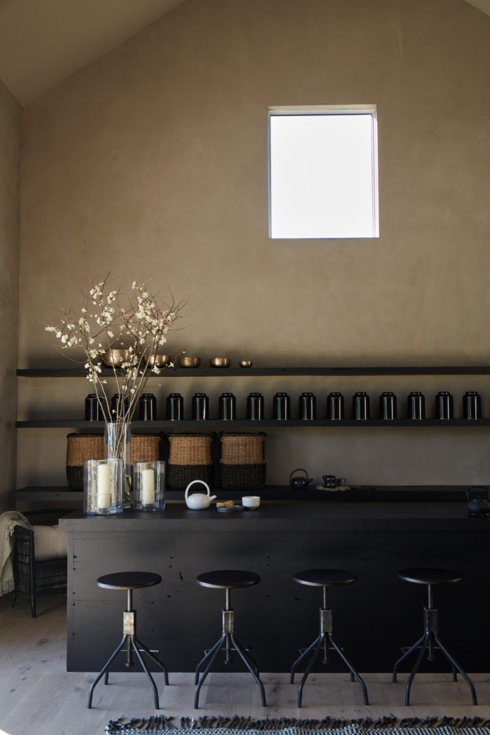 Minimalist kitchen with black stools, open shelves, canisters, wicker baskets, and a vase with twig-like flowers on a dark island.