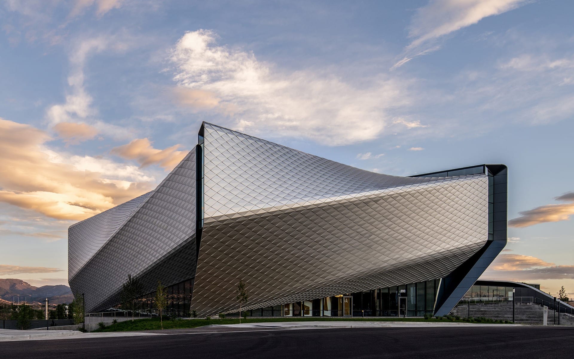 Modern angular building with reflective, textured metal facade against a sky with scattered clouds and a mountain backdrop.