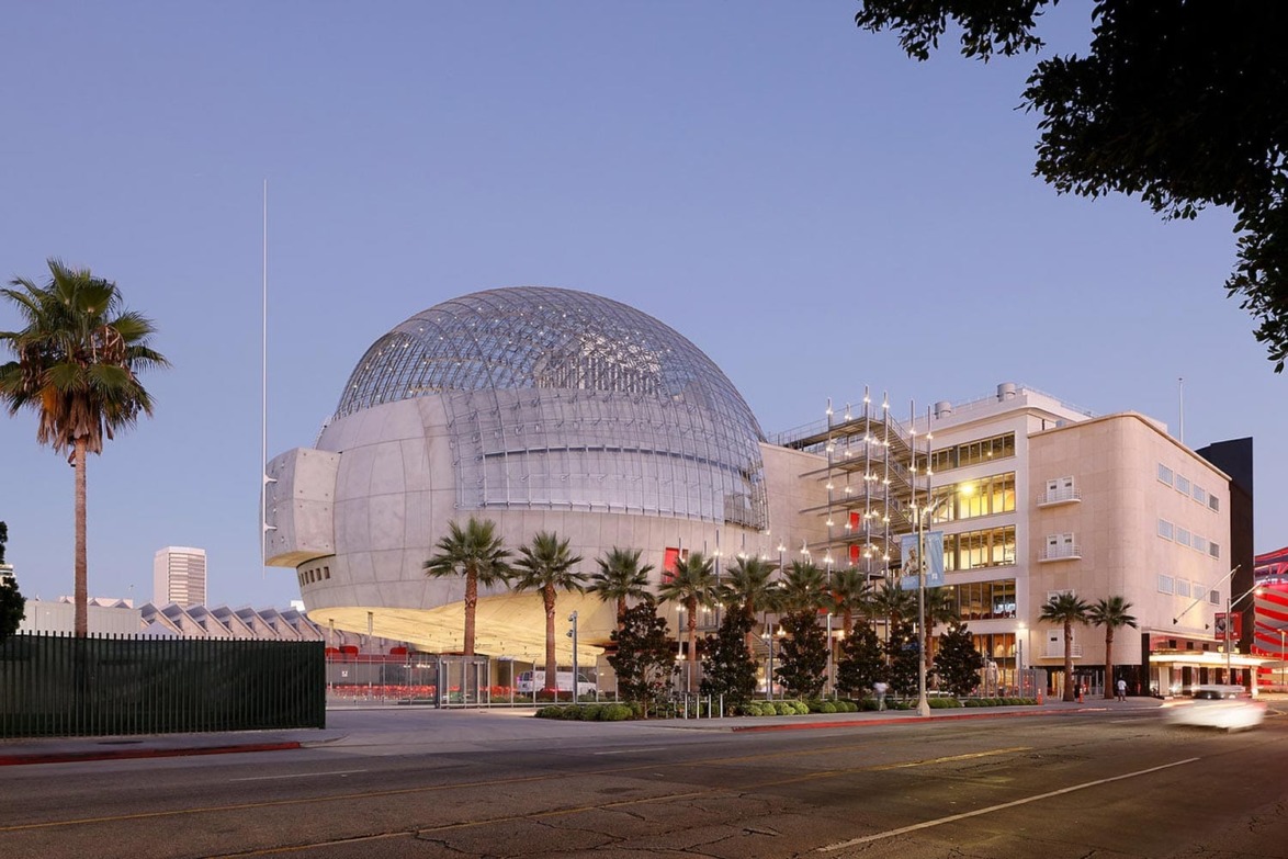 Modern architectural building with a large dome, surrounded by palm trees, viewed from the street in the evening light.
