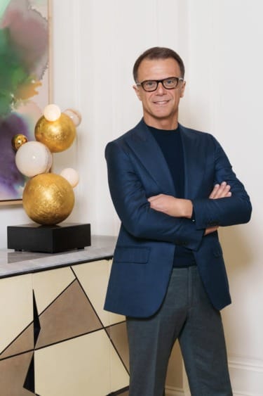 Person in a blue blazer and glasses standing next to modern art sculpture on a geometric cabinet in a stylish room.
