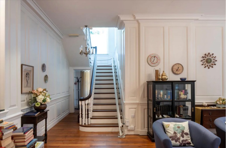 Elegant living room with a white staircase, bookcase, and wall decor, featuring wooden floors and soft lighting.