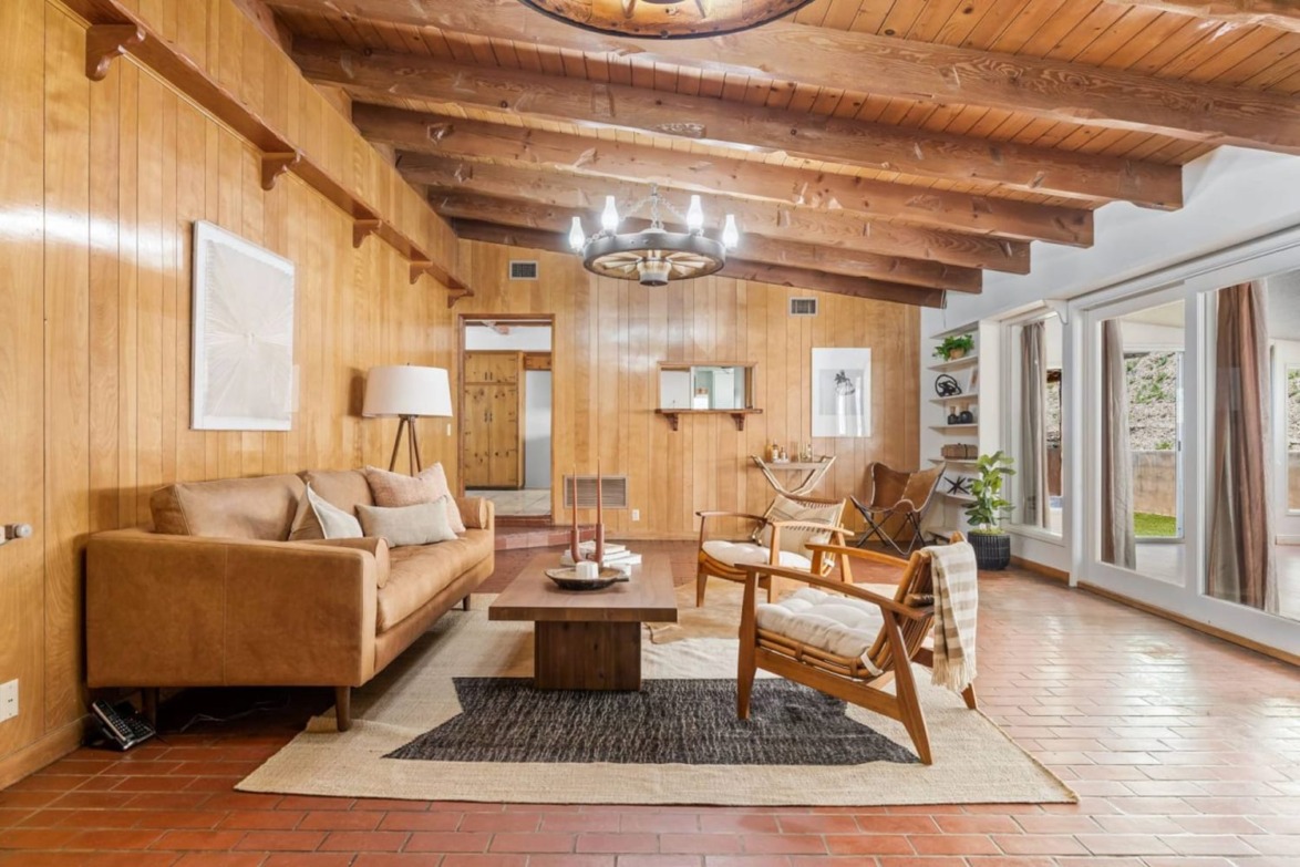Cozy living room with wooden walls, beamed ceiling, modern furniture, and large windows showing outdoor greenery.