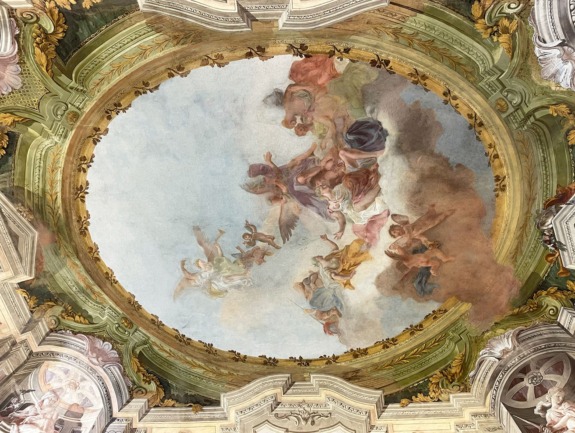 Baroque-style ceiling fresco depicting a celestial scene with angels and clouds surrounded by ornate architectural details.