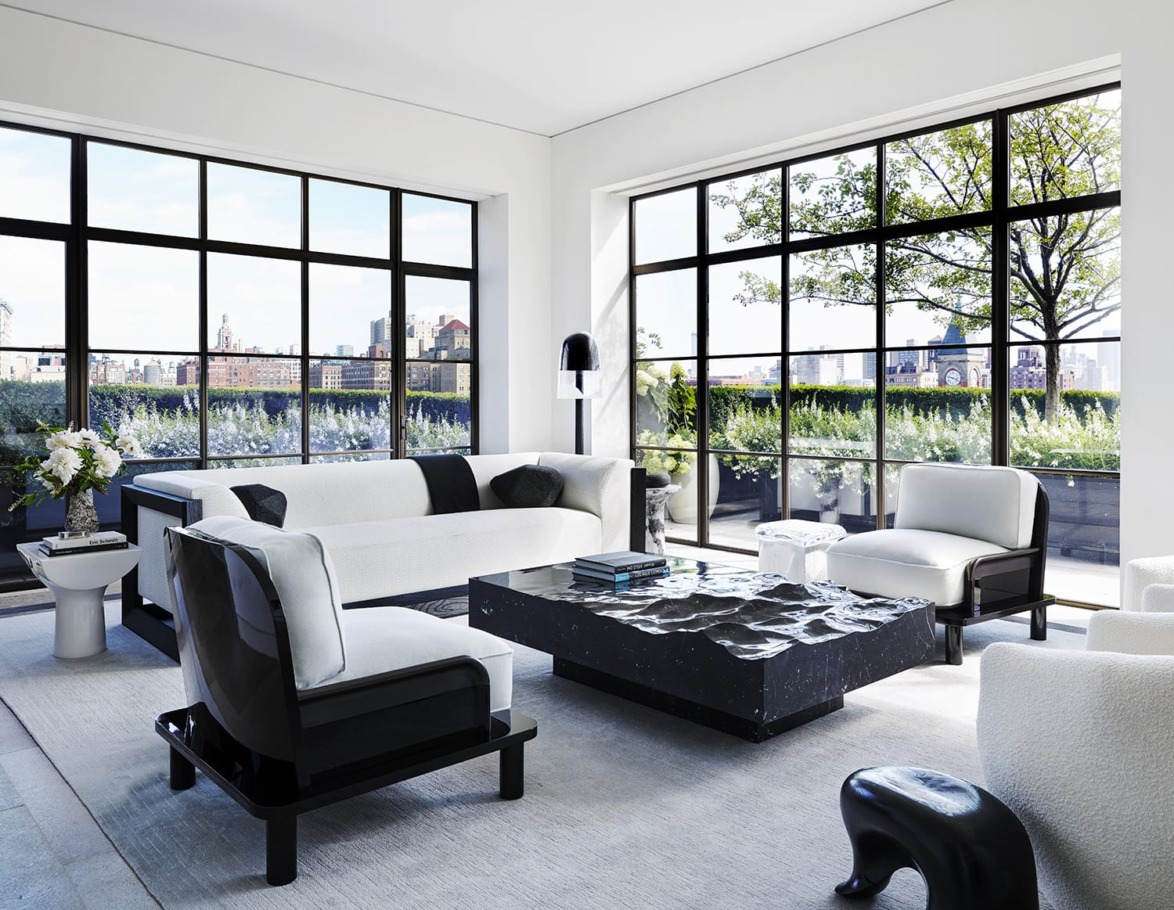 Modern living room with white and black furniture, large windows, and a view of a cityscape and greenery outside.