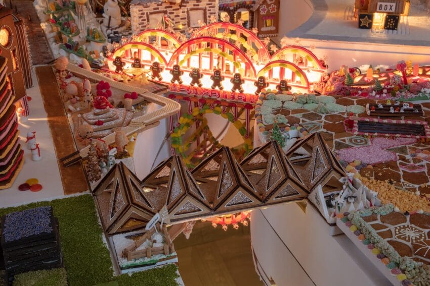 Festive gingerbread scene with colorful icing, cookie bridges, and candy decorations illuminated by warm lights.