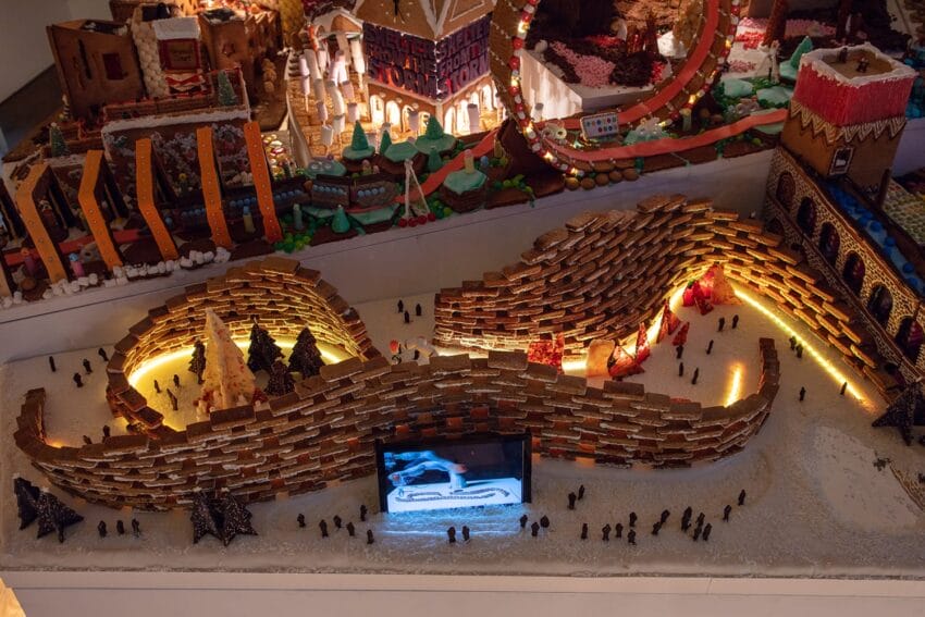 Intricately designed gingerbread sculpture featuring winding paths, trees, and miniature figures, illuminated and festive.