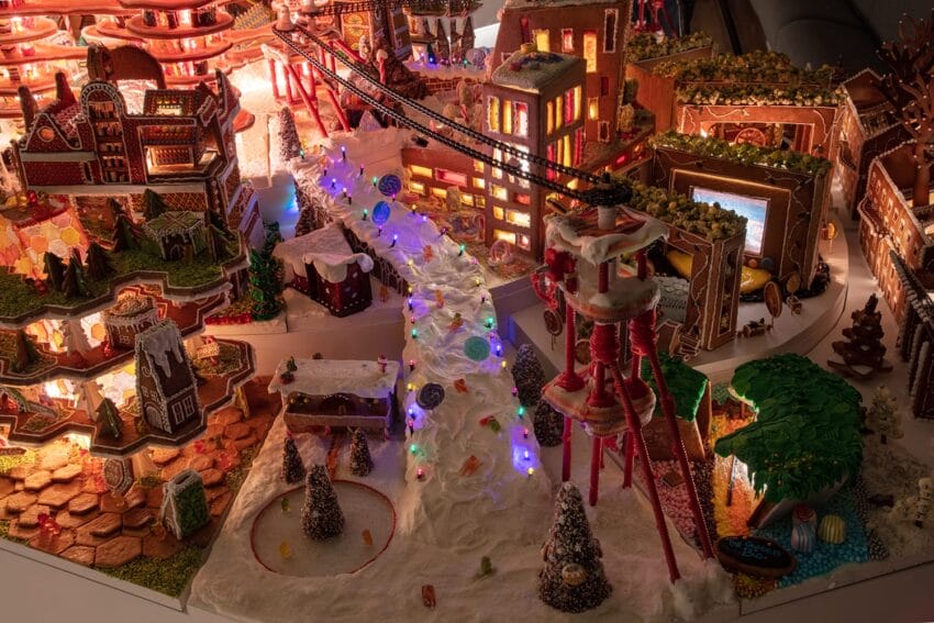 Gingerbread village with illuminated buildings, snowy paths, trees, and colorful decorations creating a festive scene.