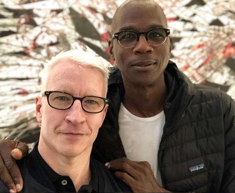 Two people wearing glasses and casual clothing standing together, with a patterned background behind them.