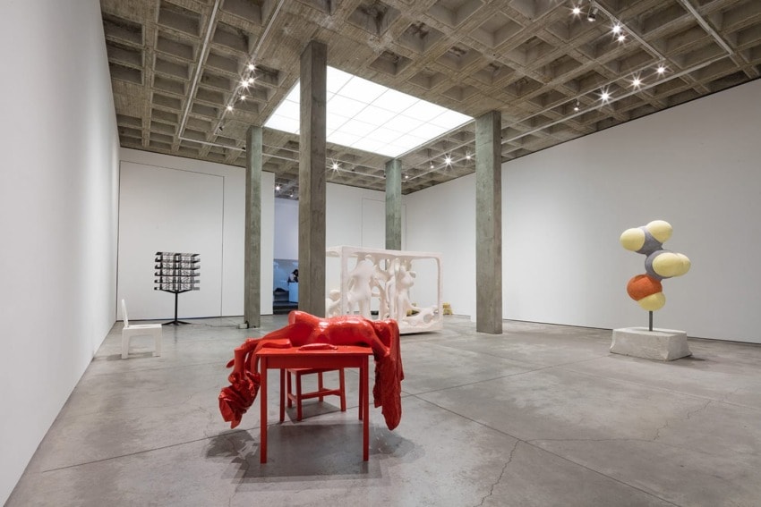 Modern art gallery with various sculptures displayed in a spacious room with concrete walls and a large skylight.