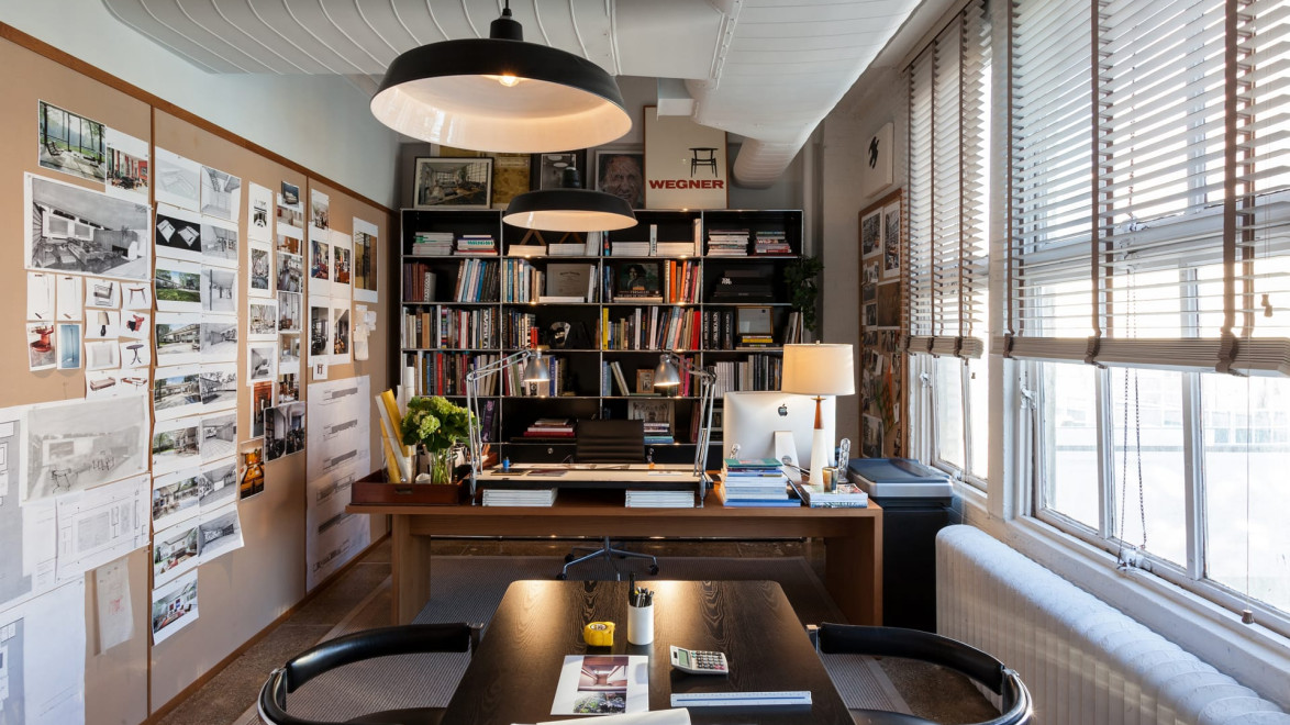 Modern office with bookshelves, desk, and wall art, featuring large windows with blinds and overhead industrial-style lights.