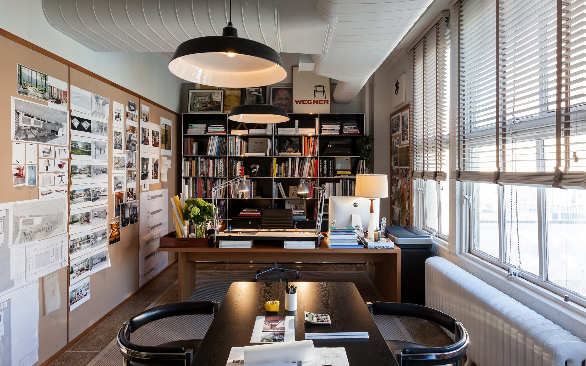 Modern office with bookshelves, desk, and wall art, featuring large windows with blinds and overhead industrial-style lights.