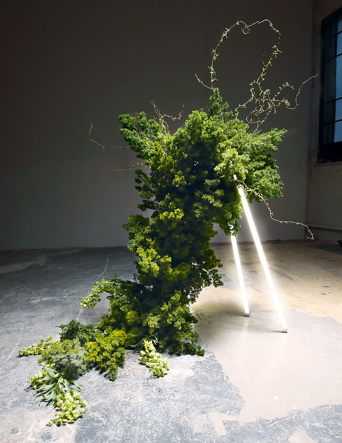 Abstract green plant sculpture with branches and leaves, supported by neon tubes, placed on a concrete floor in a dim room.