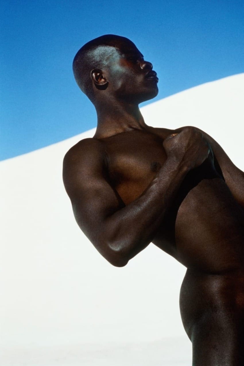 Person posing against a backdrop of white sand dunes and a clear blue sky, showcasing strong, defined muscles and confident posture.