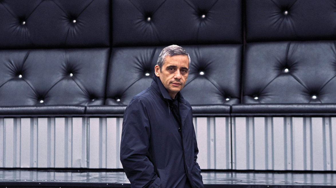 Man in a dark coat standing in front of a tufted black wall outdoors.