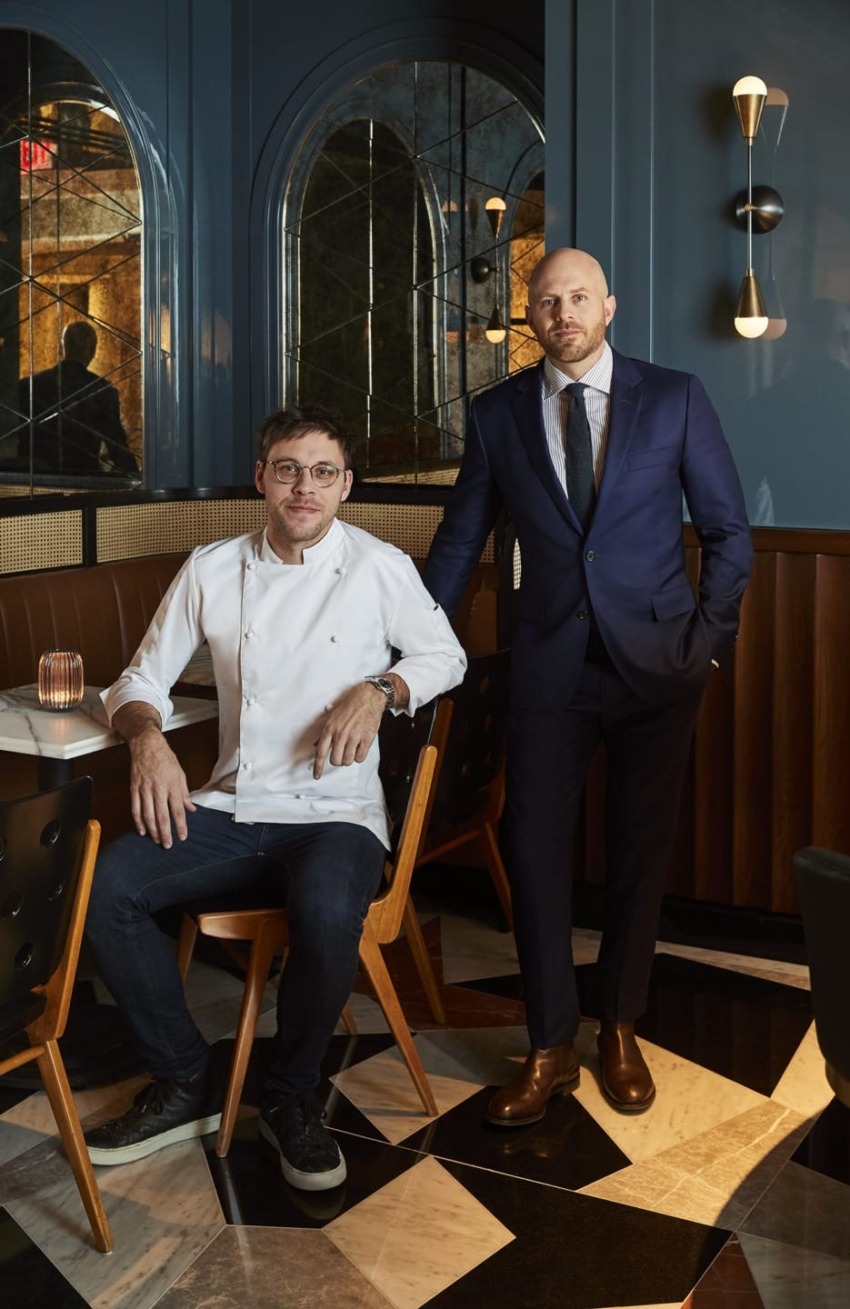Two men in a stylish restaurant setting, one seated in a chef's coat, the other standing in a suit.