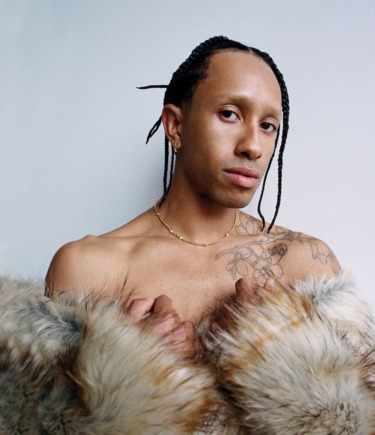 Person with braided hair and tattoos wearing a fur coat against a plain background.