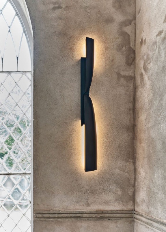 Modern black wall sconce emitting soft light against textured beige wall near an arched window with a diamond pattern.